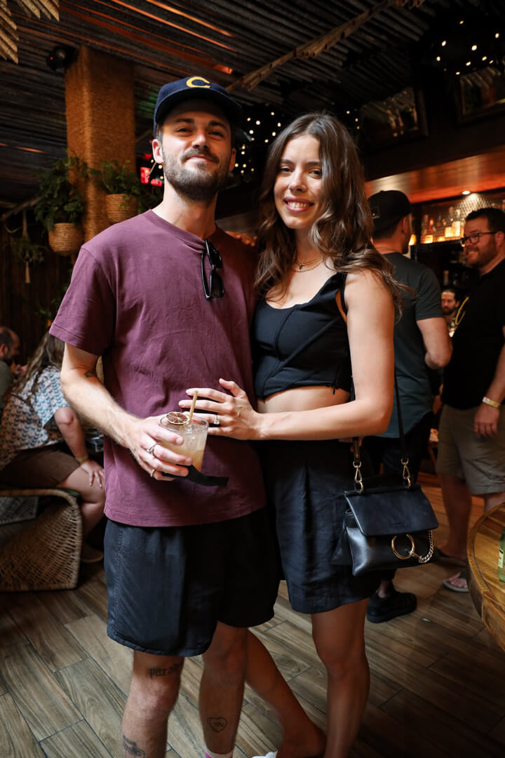 A man and woman standing and smiling for the camera. the man is holding a cocktail in his hand